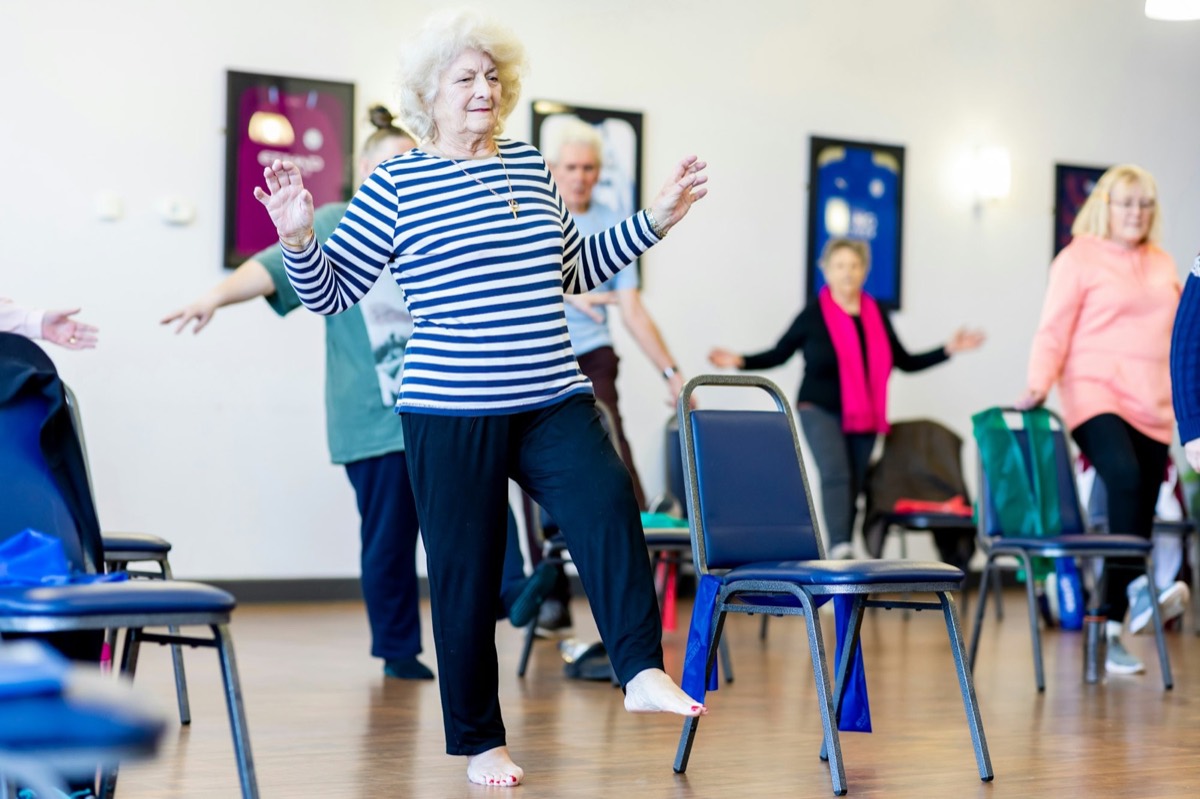 A group of older women in light clothing smile and stretch their arms together in a bright exercise class. Stock photograph of models; not a real place.