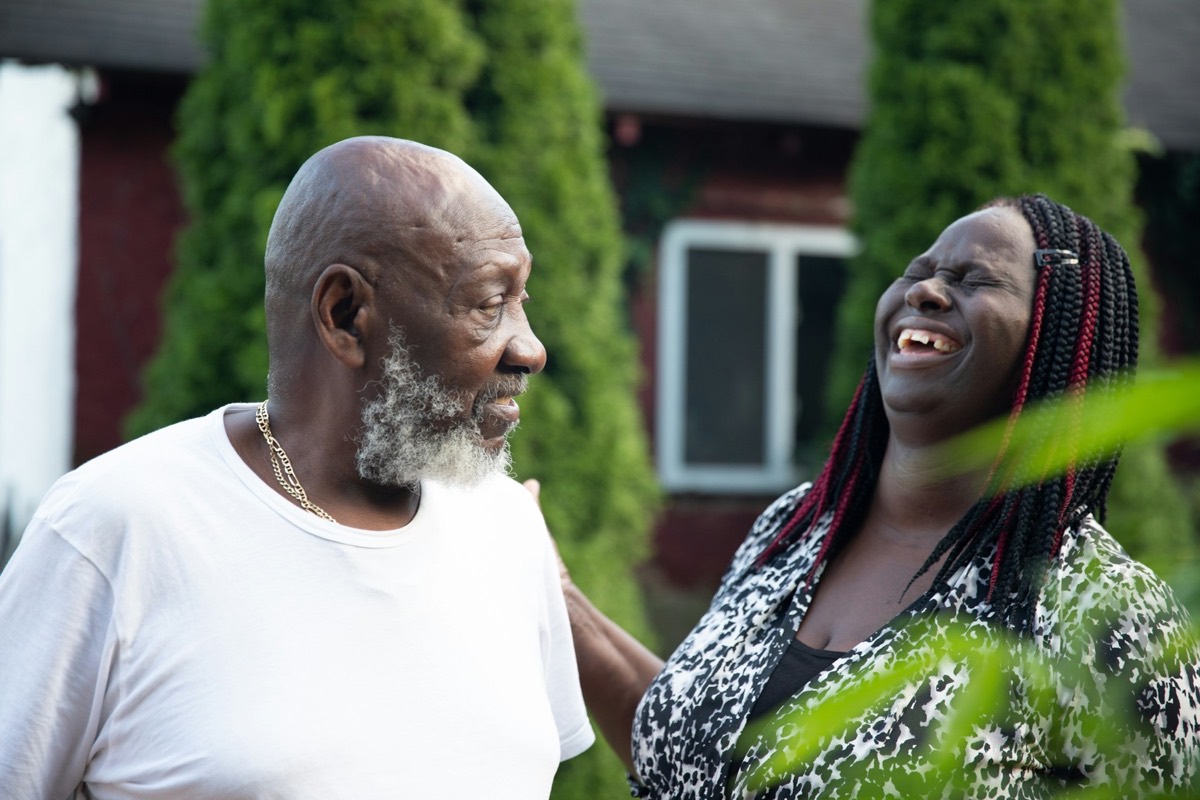 An older Black man with a grey beard and an older Black woman with long braids laugh together in a garden. Stock photograph of models; not a real community or resident.