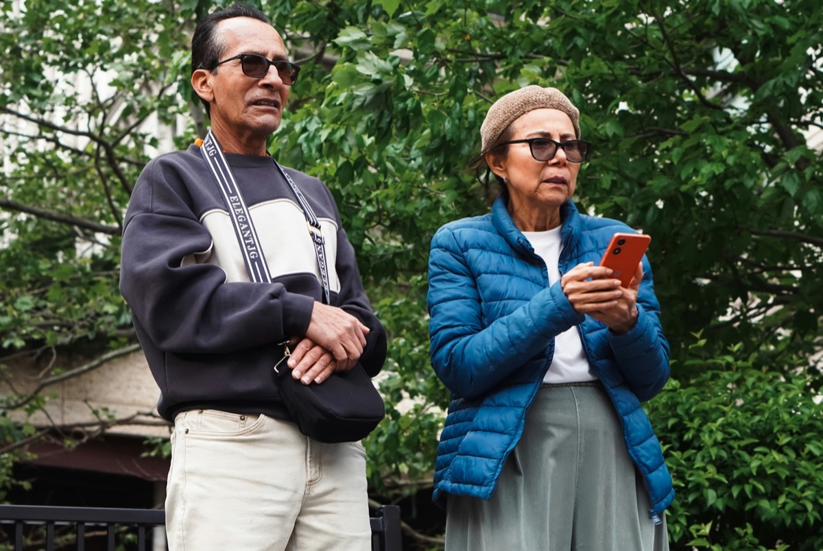 An older man and woman in sunglasses stand together outdoors among trees; the woman holds a red phone. Stock photograph of models; not a real place.