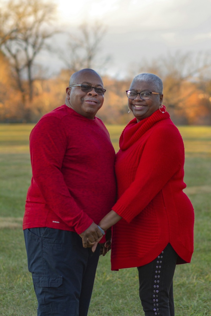 An older Black couple in matching red tops stand close together on a lawn at sunset, holding hands. Stock photograph of models; not a real place.