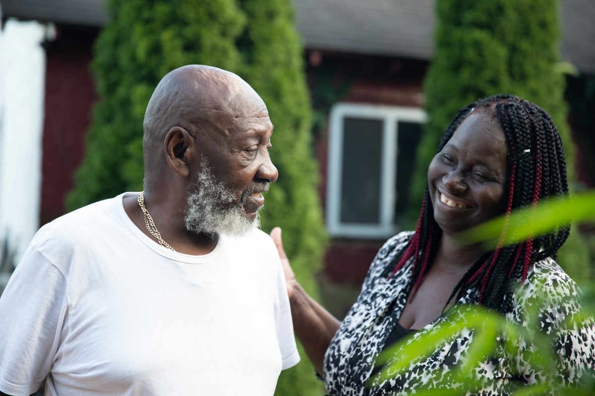 An older Black man with a grey beard and an older Black woman with long braids smile as they sit close together outdoors. Stock photograph of models; not a real place.