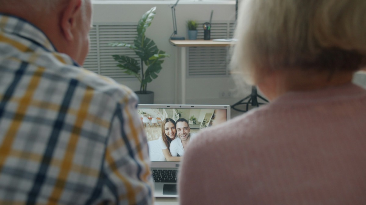Two older adults, seen from behind, sit at a laptop and talk by video with a smiling younger couple on the screen. Stock photograph of models; not a real place.